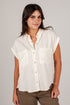 A woman wearing a short-sleeve button-up shirt in off-white with two chest pockets, paired with brown pants, against a plain light gray studio background.