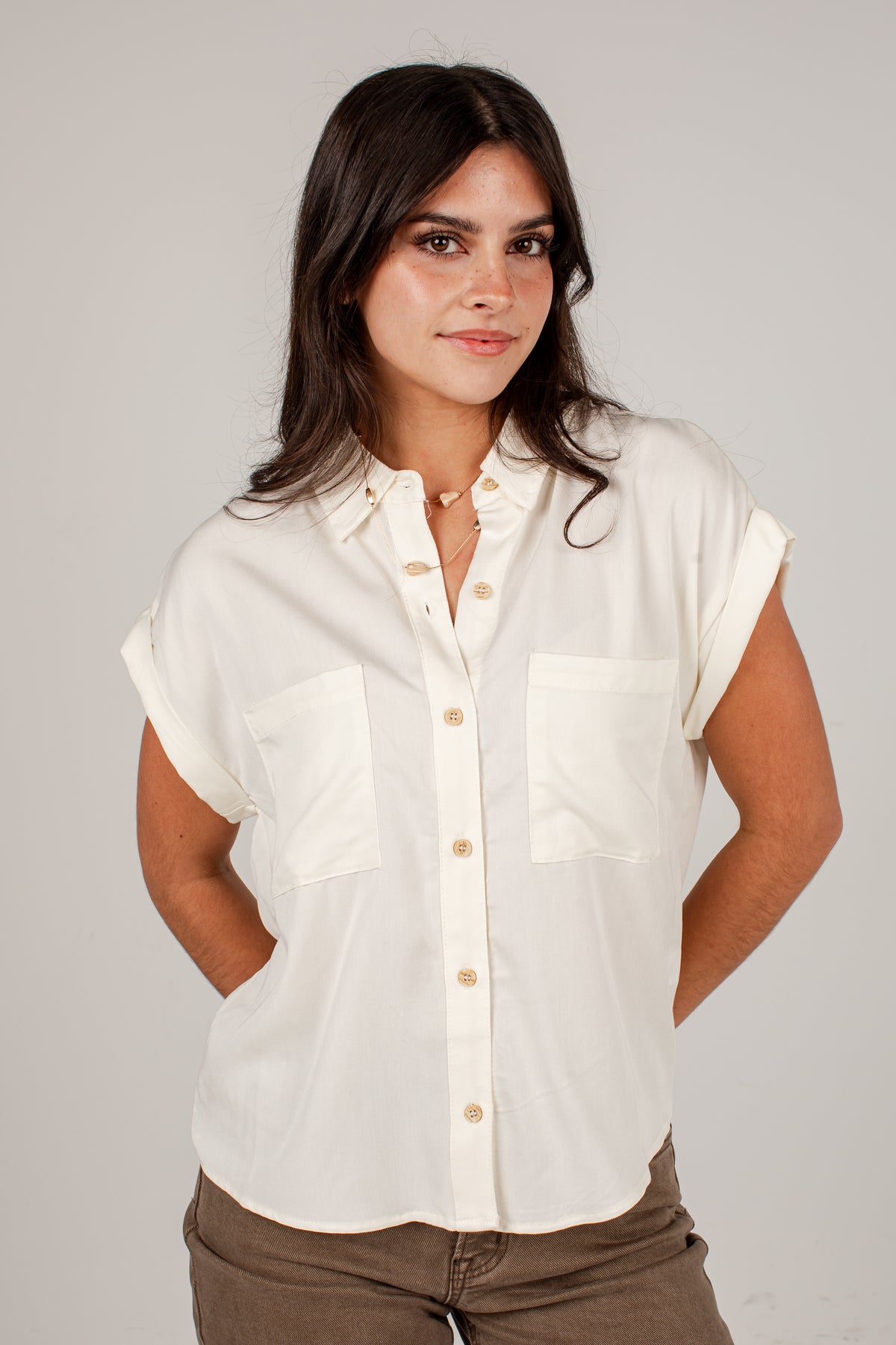 A woman wearing a short-sleeve button-up shirt in off-white with two chest pockets and tan buttons, paired with brown pants, against a plain light gray studio background.