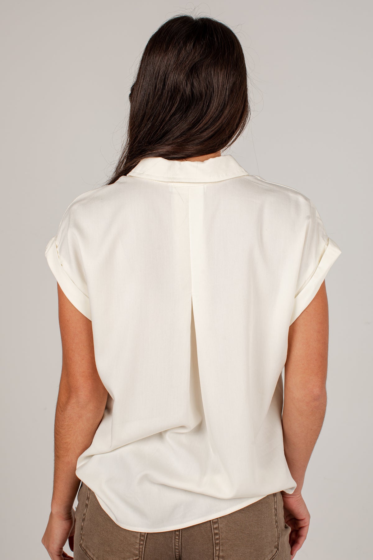 A woman viewed from the back wearing a short-sleeve cream-colored blouse and brown pants, set against a plain light gray studio background.