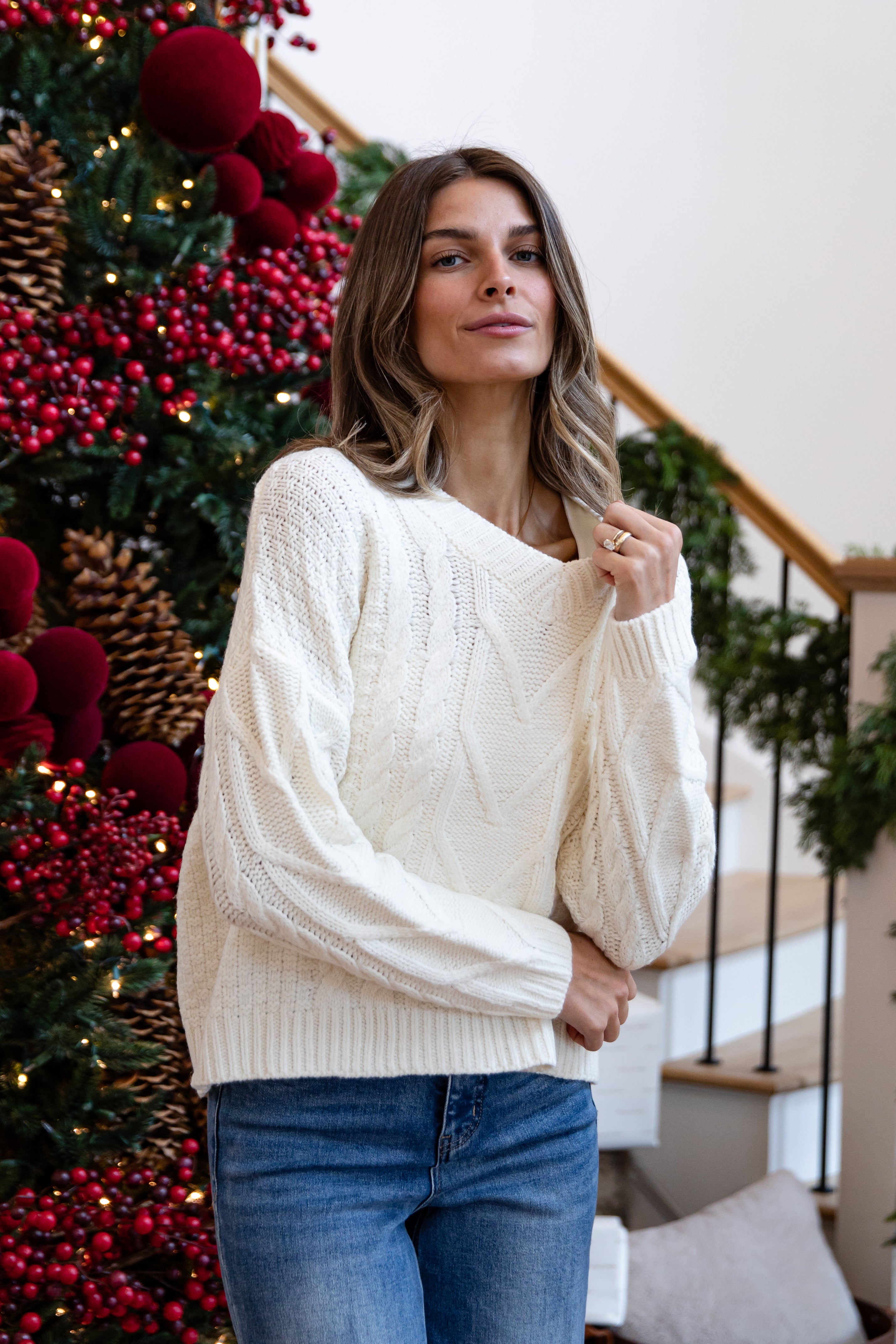 Woman wearing a cream sweater and blue jeans standing in front of a decorated Christmas tree.