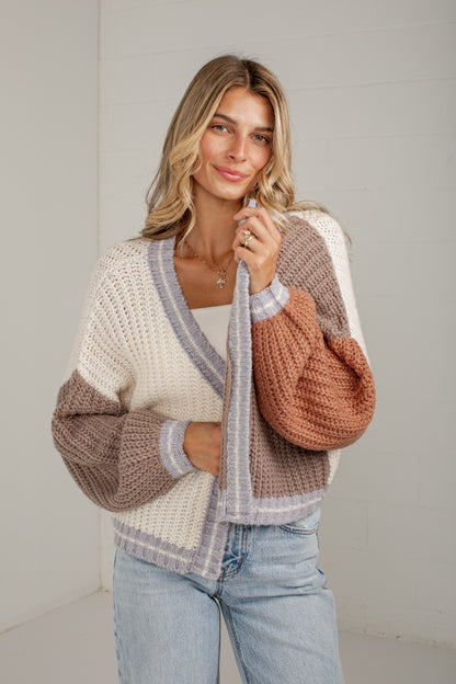 A woman wearing a chunky knit color-block cardigan with cream, taupe, and terracotta sections, paired with light-wash blue jeans, standing against a light gray wall.