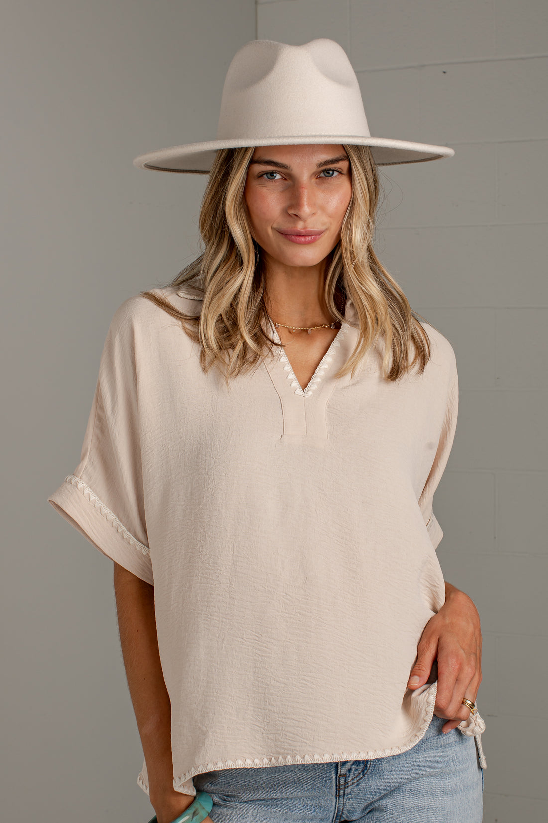 A woman wearing a cream-colored short-sleeve top with a split V-neck and tie details, paired with a matching wide-brim hat and light blue jeans, standing against a gray paneled wall.