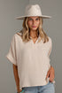 A woman wearing a cream-colored short-sleeve top with a split V-neck and tie details, paired with a matching wide-brim hat and light blue jeans, standing against a gray paneled wall.