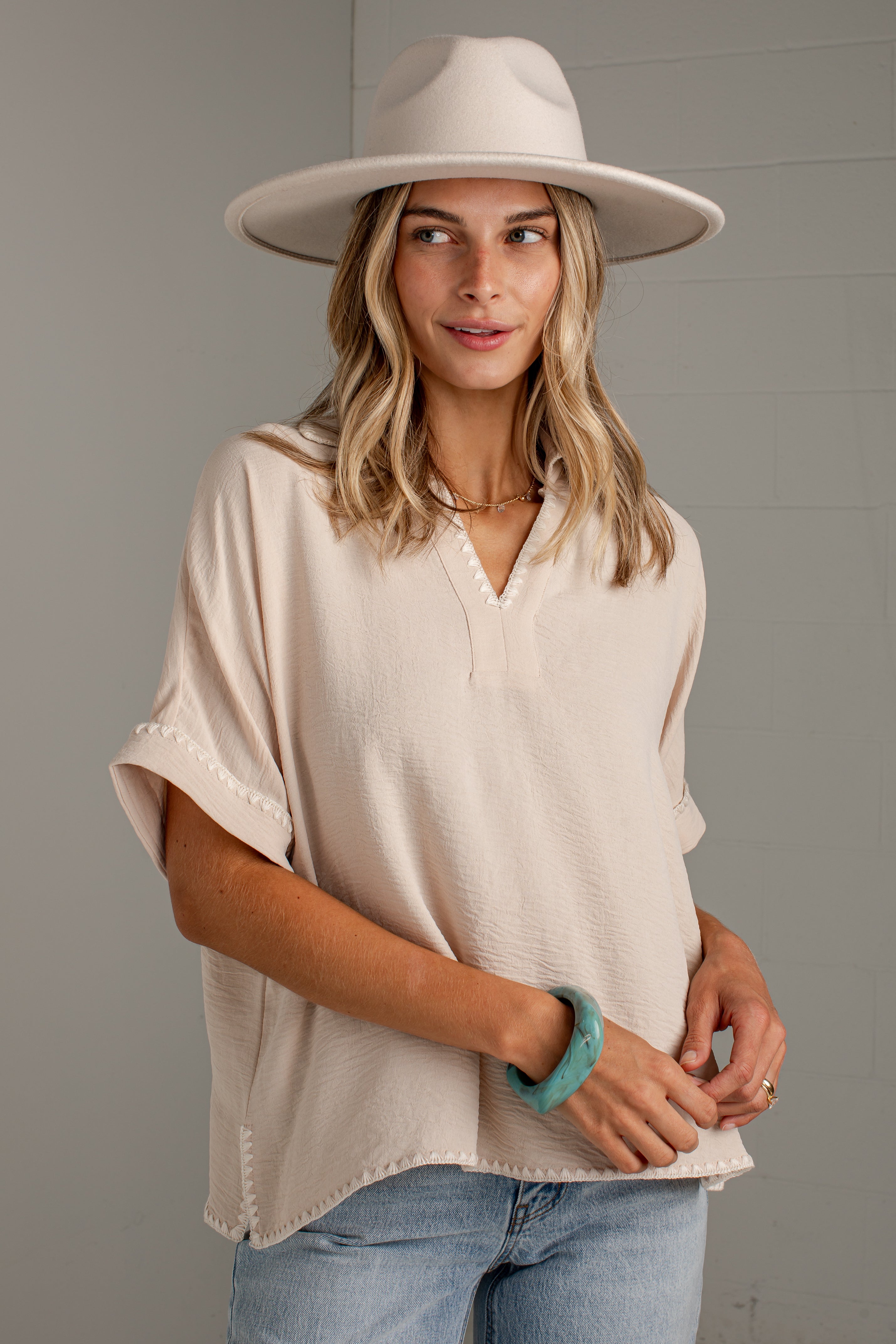 A woman wearing a light beige short-sleeve blouse with a split V-neck and scalloped edges, paired with light blue jeans and a wide-brimmed cream hat, standing against a gray wall.