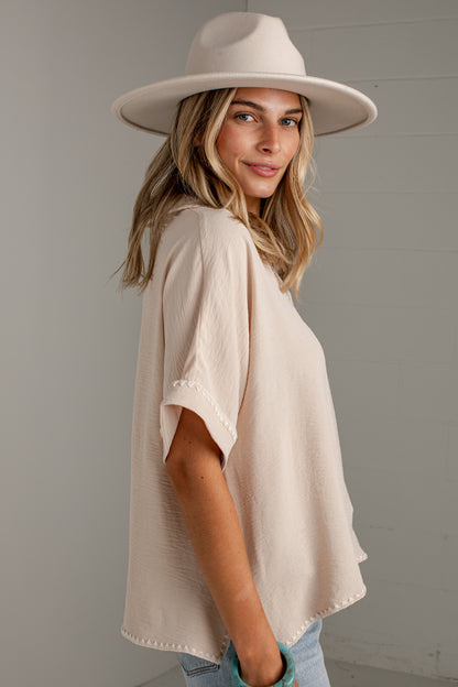 A woman wearing a cream-colored, short-sleeve textured top and a matching wide-brim fedora hat, photographed against a light gray paneled wall.