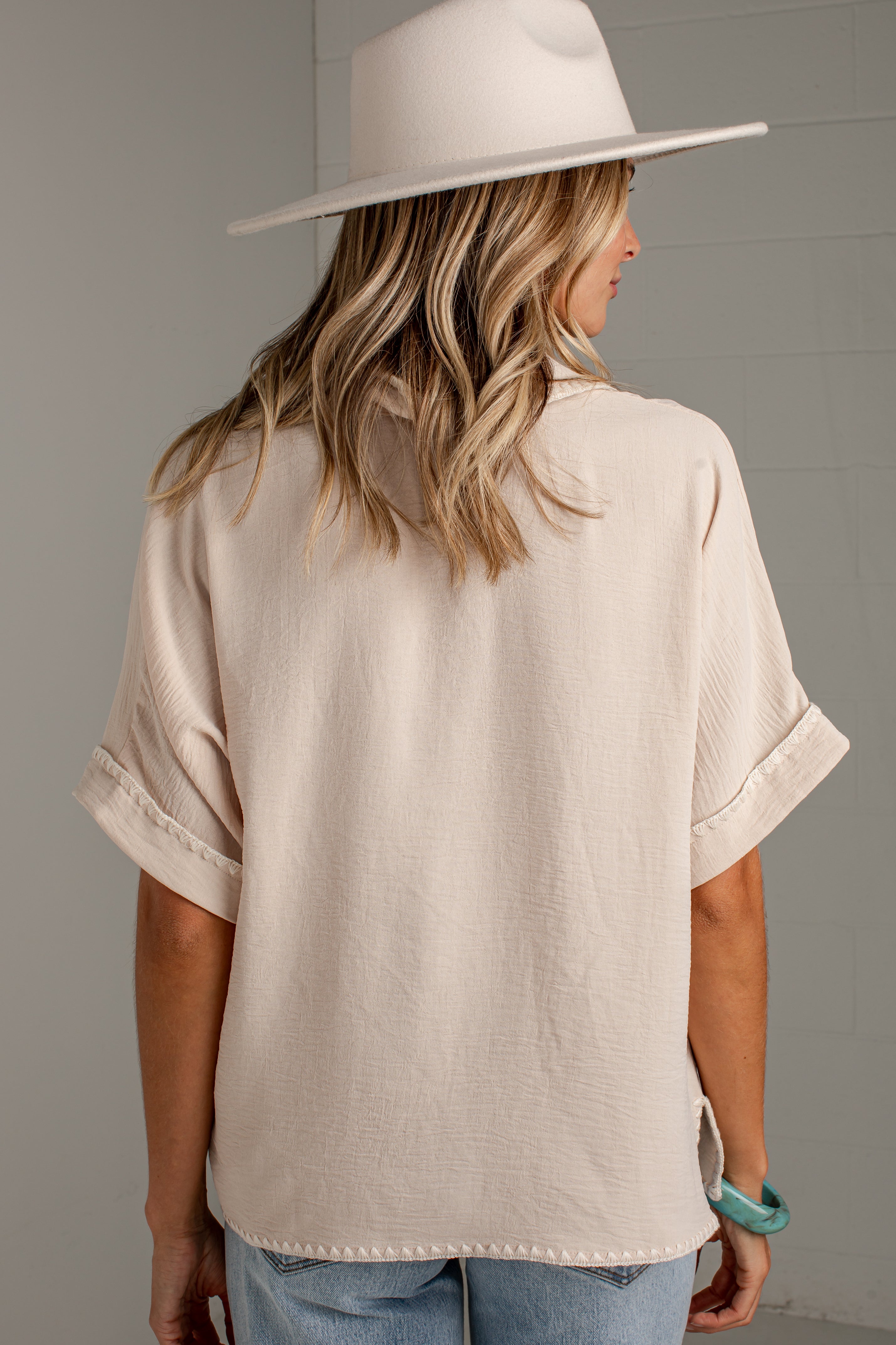 A woman shown from the back wearing a cream-colored short-sleeve top with rolled cuffs, paired with light blue jeans and a matching cream wide-brim hat, set against a neutral gray background.