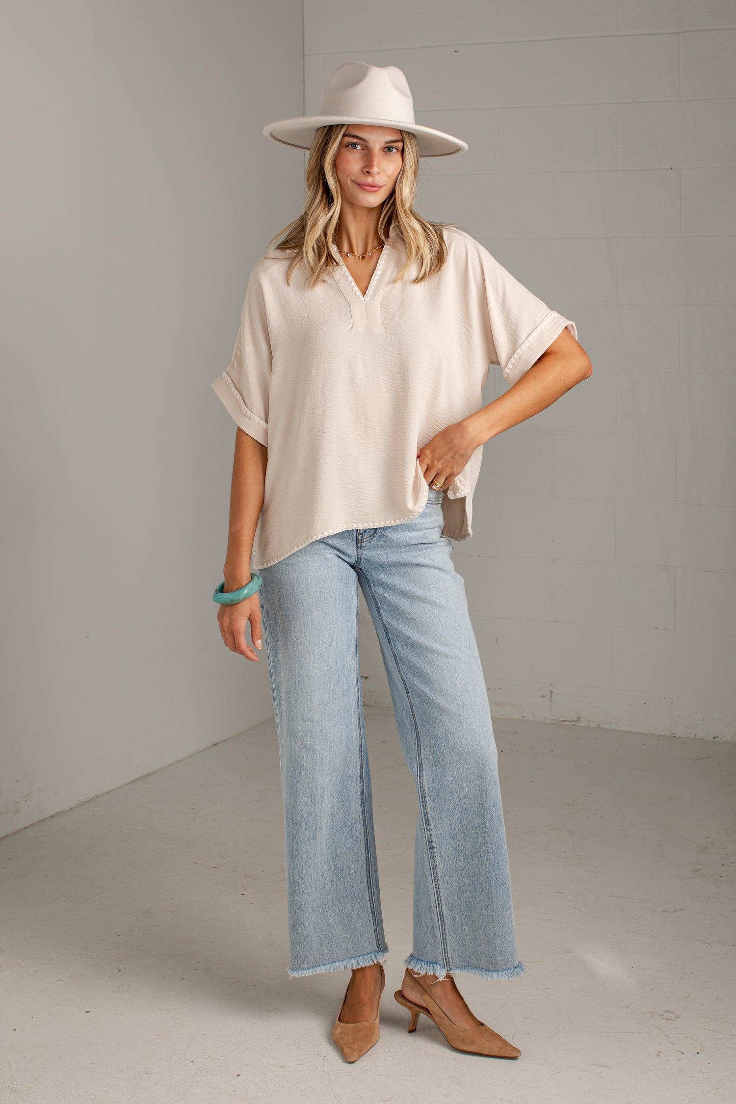 A woman modeling a cream short-sleeve collared blouse, light-wash wide-leg jeans, a cream wide-brim hat, and tan heeled mules in a bright studio setting.