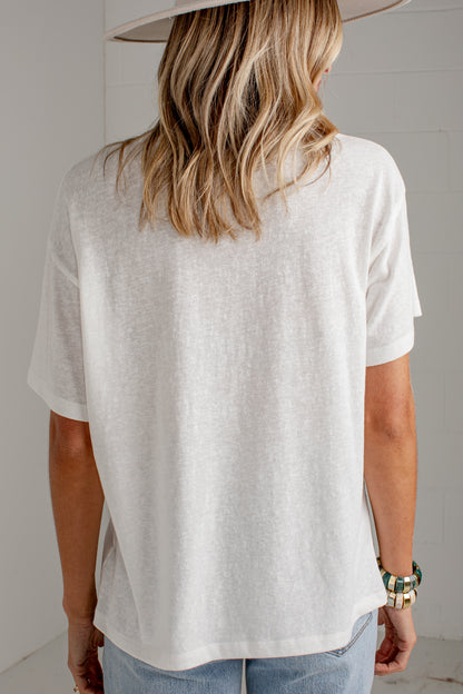 A woman viewed from behind wearing an oversized off-white short-sleeve t-shirt, light blue jeans, and a wide-brimmed light-colored hat, set against a neutral light gray background.
