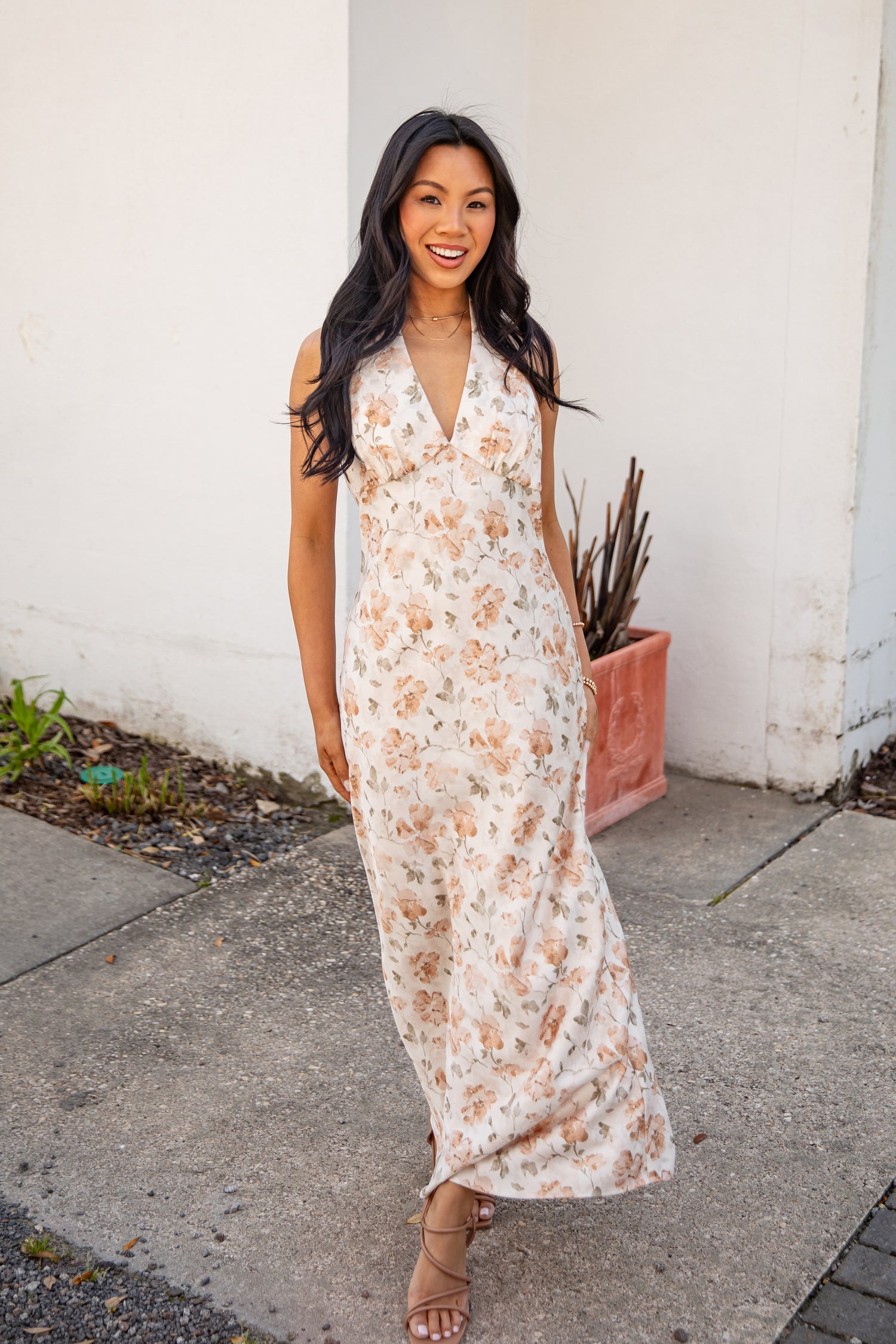 Woman in a floral dress standing on a sidewalk with a white wall and plant in the background