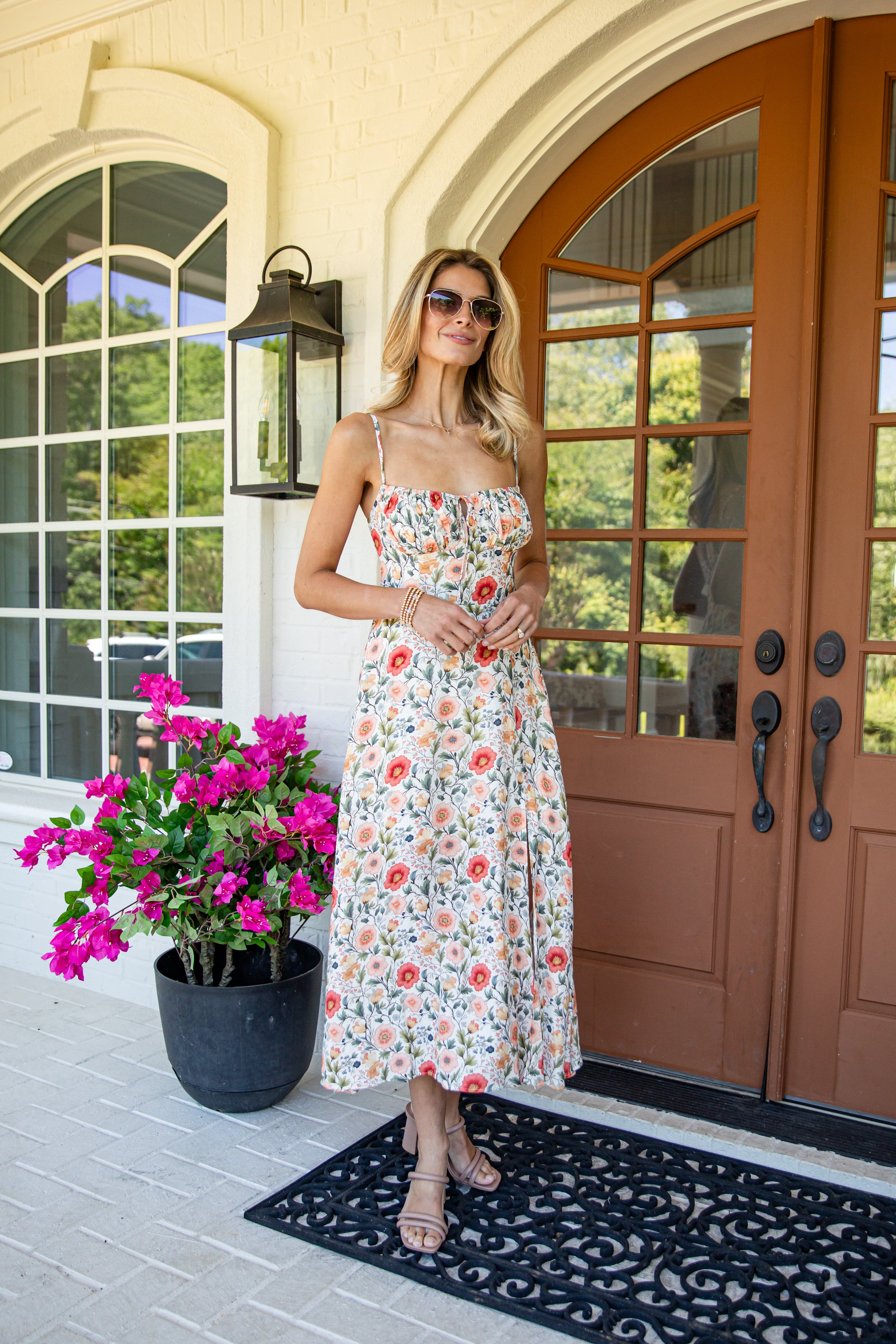 A woman wearing a floral spaghetti-strap midi dress and sunglasses stands on a porch in front of brown double doors, with a potted pink flower on the left.