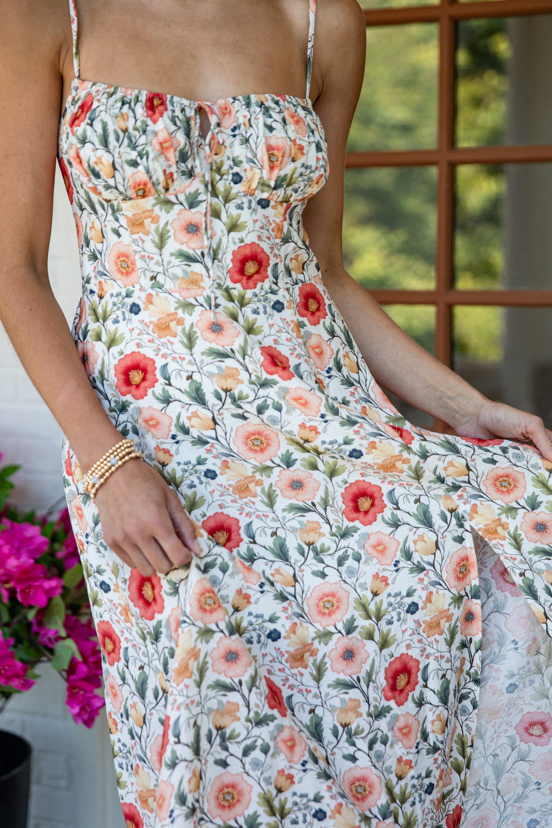 A woman wearing a sleeveless white maxi dress with a colorful floral print in red, orange, pink, and green, standing in front of a window.