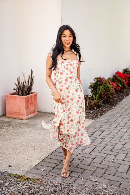 Woman in a floral dress standing on a sidewalk with plants in the background