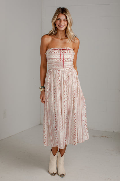 A woman modeling a strapless midi dress with a pink and white bohemian pattern, standing in a white studio setting.