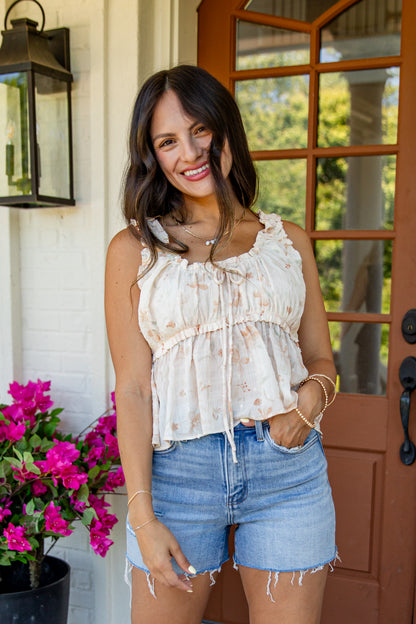 A woman wearing a cream-colored sleeveless top with a subtle floral pattern and ruffled tie details, paired with light-wash denim shorts, standing on a porch in front of a brown glass door.