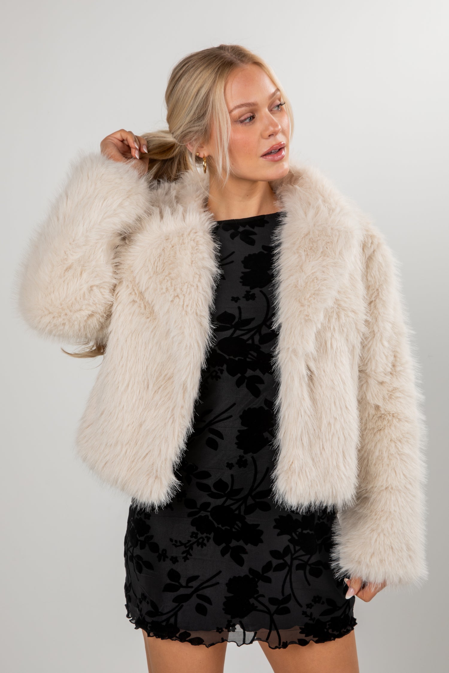 Woman wearing a black dress with floral patterns and a beige fur coat against a plain background