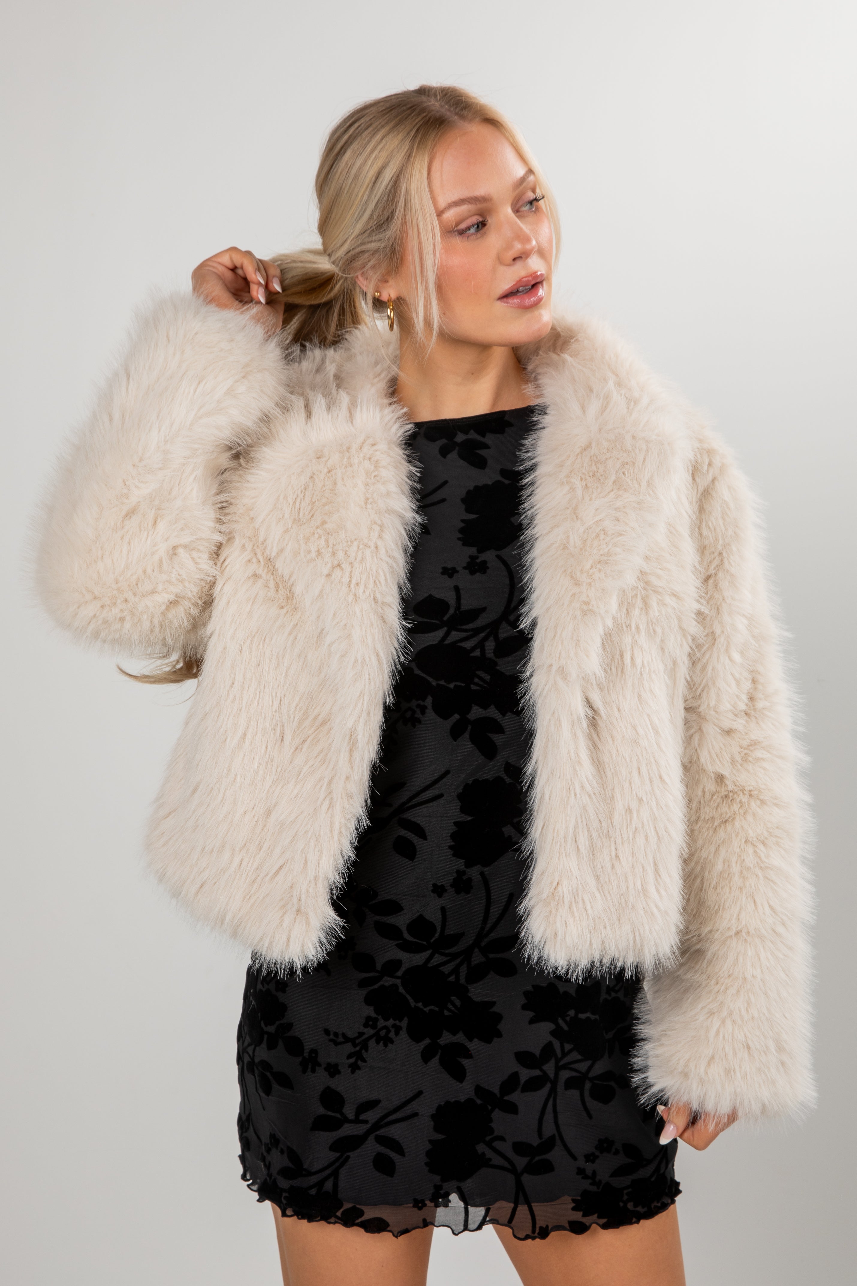 Woman wearing a black dress with floral patterns and a beige fur coat against a plain background