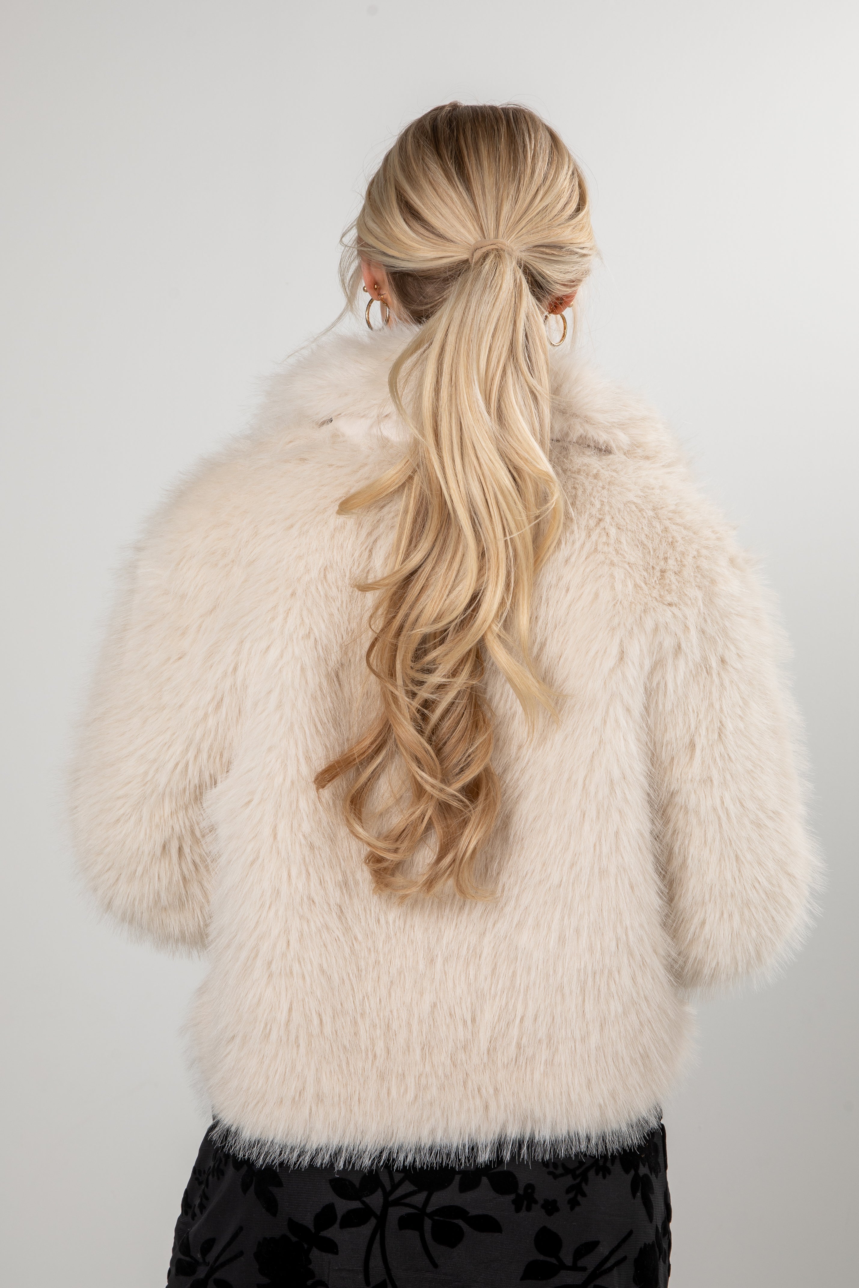 Person wearing a fluffy white coat with long blonde hair styled in a ponytail against a plain background