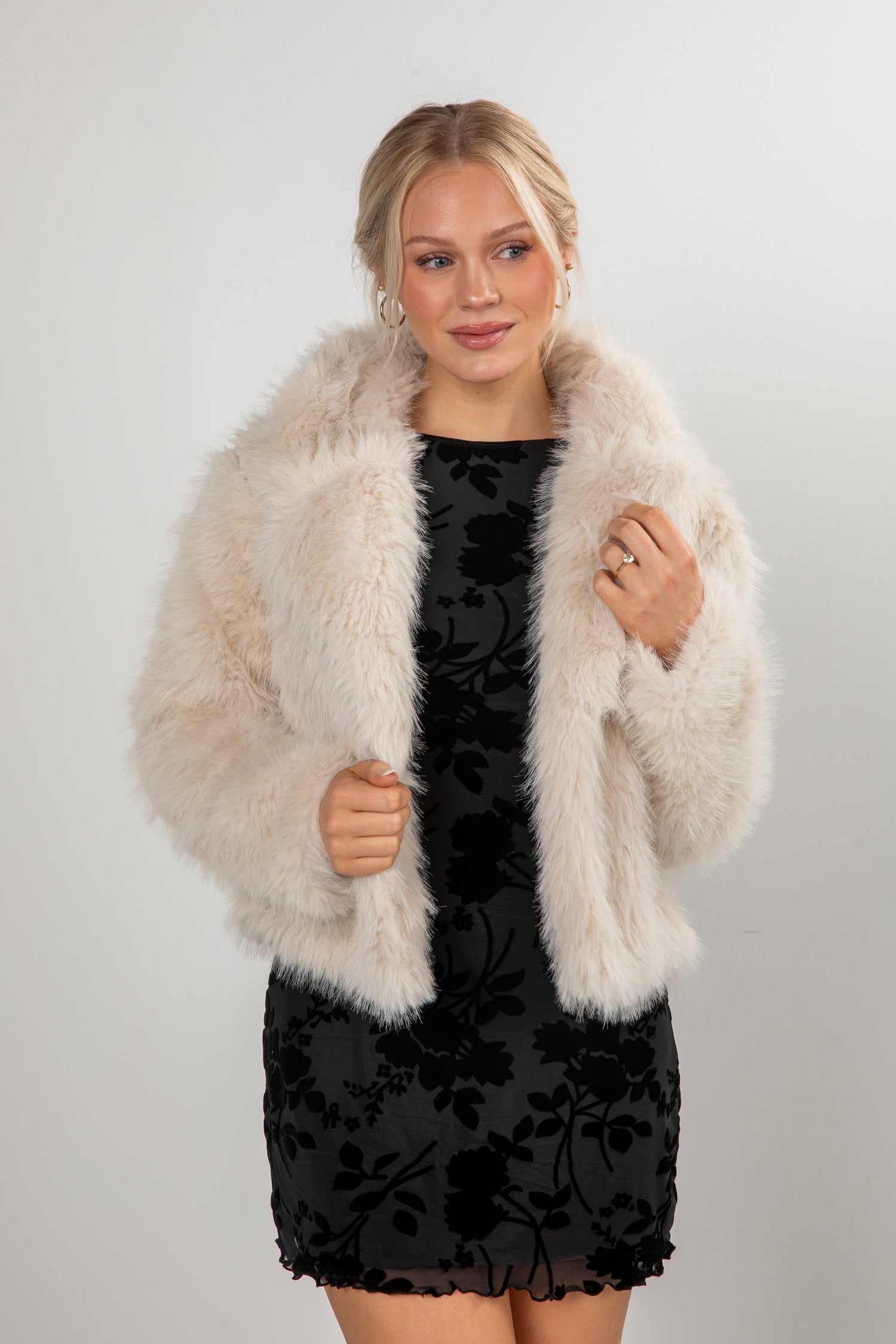 Woman wearing a black dress with floral patterns and a beige fur coat against a white background