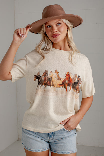 A woman wearing a cream-colored short-sleeve t-shirt with a graphic print of a herd of horses, paired with denim shorts and a brown wide-brim hat, posing against a light gray wall.