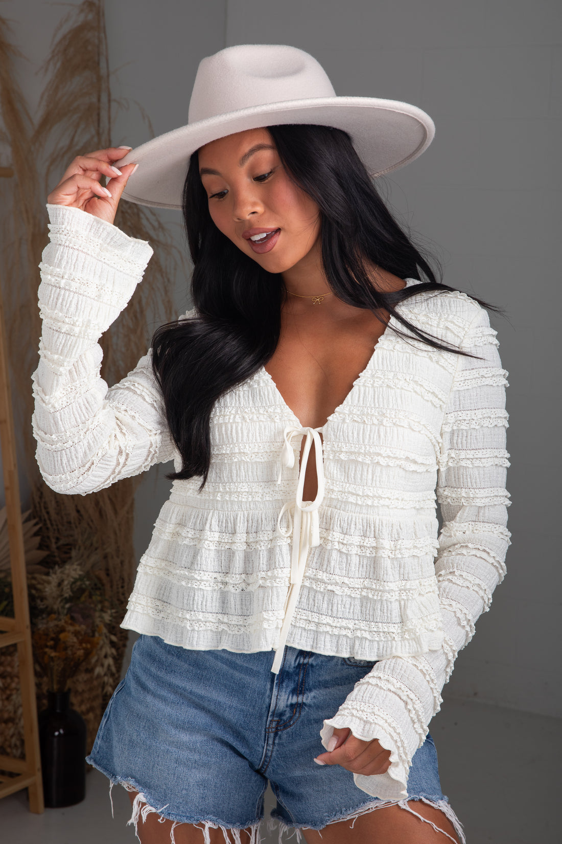 Woman wearing a white lace top and denim shorts with a white hat indoors.