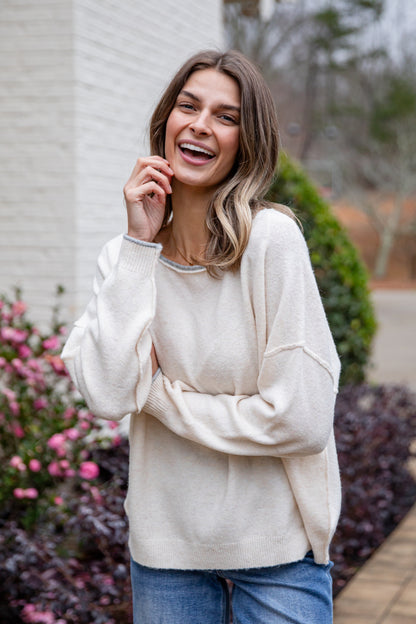 Woman wearing a white sweater and blue jeans standing outdoors with flowers and a building in the background