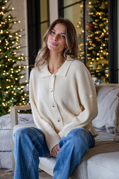 Woman wearing a cream cardigan and blue jeans sitting on a couch with Christmas trees in the background