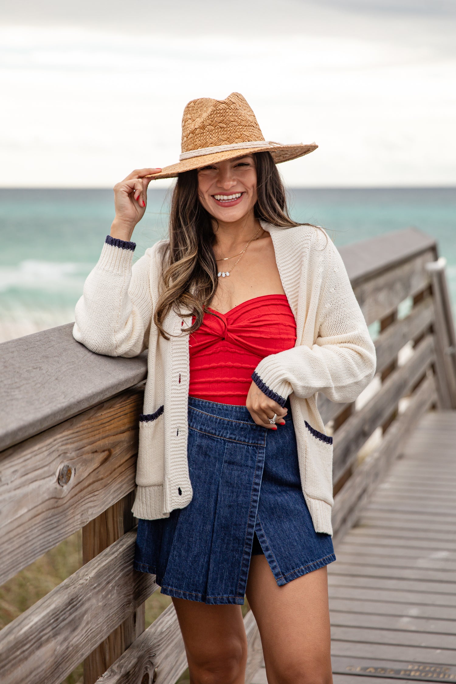 Woman wearing a red top, denim skirt, and straw hat by the ocean.