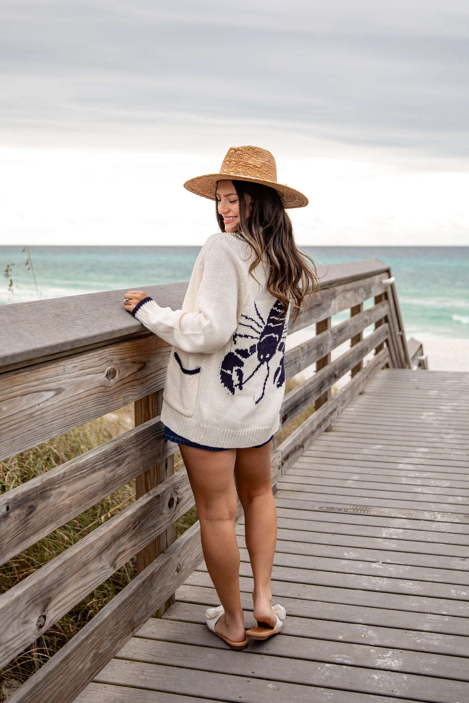 Woman standing on a wooden boardwalk by the ocean, wearing a straw hat and a patterned sweater.