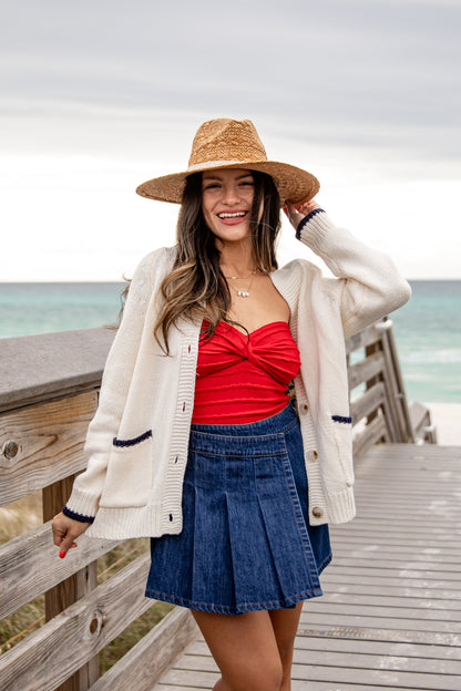 Woman in a red top and blue skirt standing on a wooden boardwalk by the ocean.