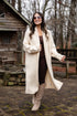 Woman in a long cream coat standing outdoors near a wooden cabin.