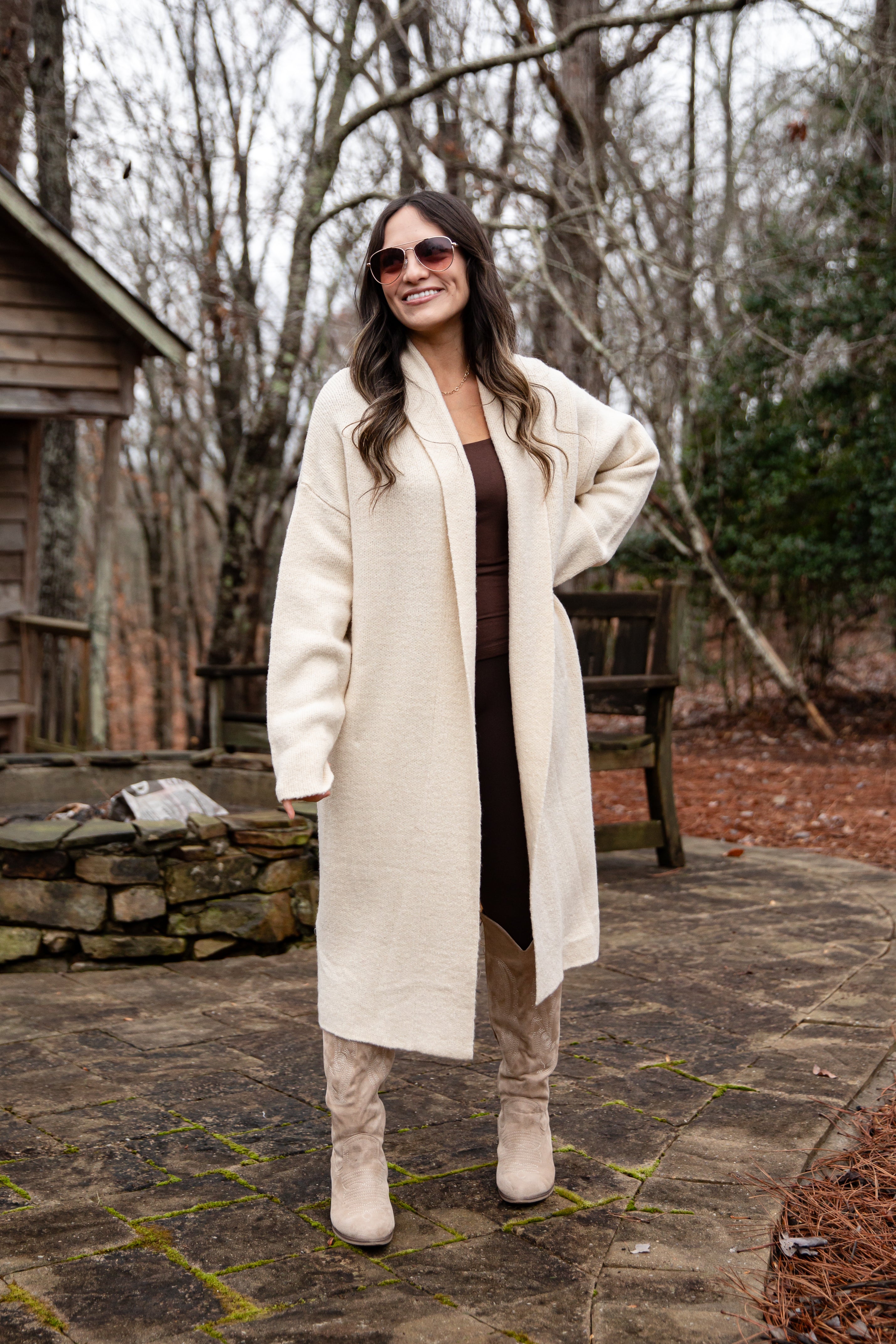 Woman wearing a long white coat and sunglasses outdoors in a natural setting