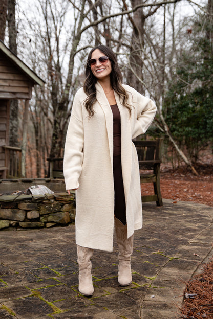 Woman wearing a long white coat and sunglasses outdoors in a natural setting