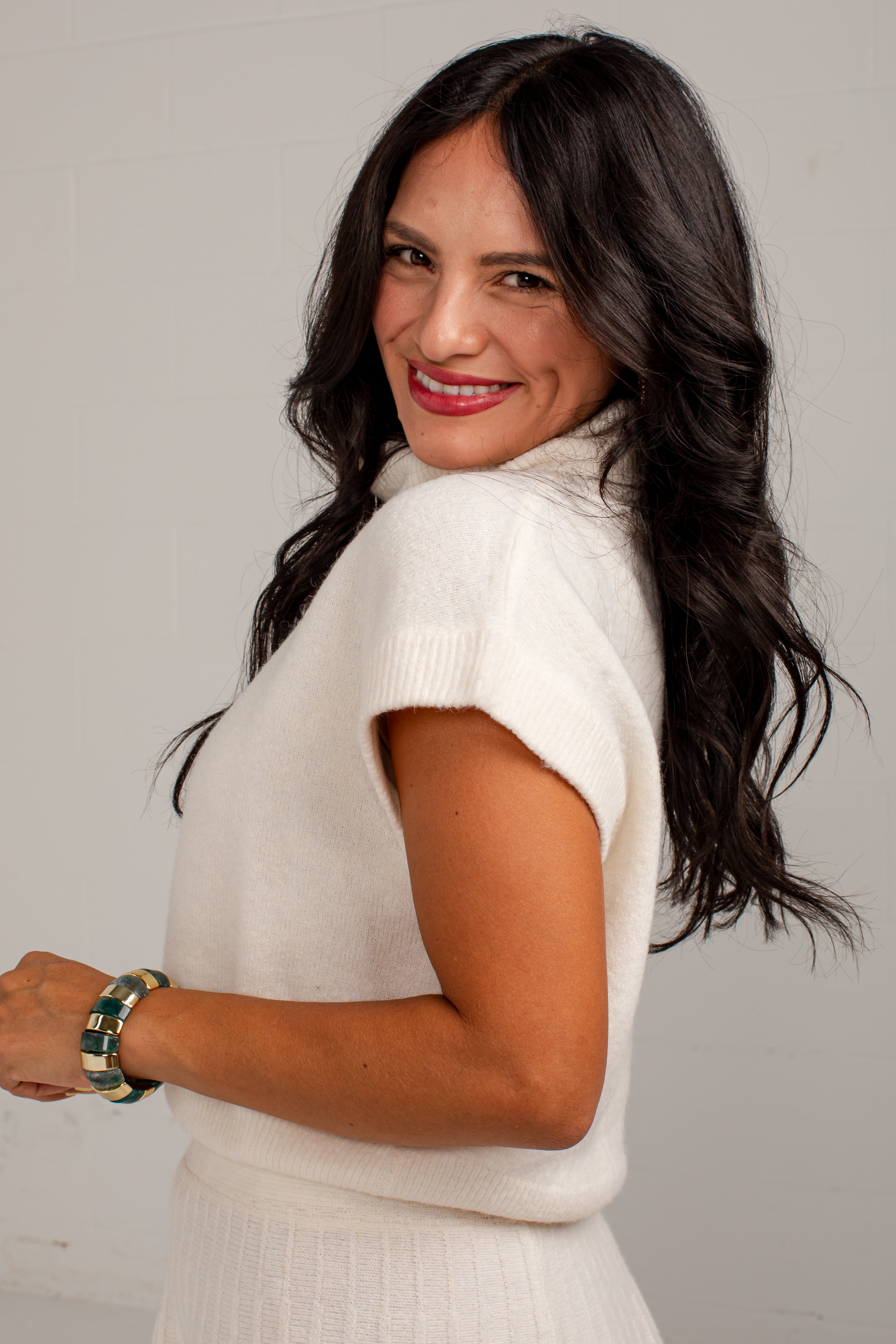 A woman with long, wavy dark hair is shown from the side and back, wearing a cream-colored short-sleeve knit sweater and matching skirt. She is wearing a green and gold beaded bracelet on her wrist.