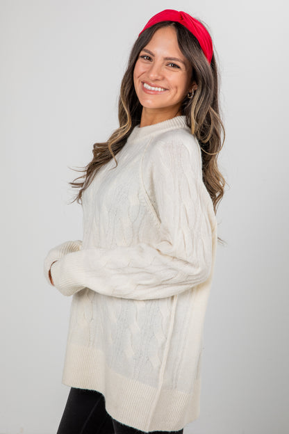 Woman wearing a white cable knit sweater and red headband against a plain background