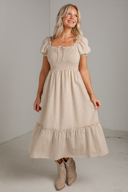 A woman modeling a beige and white gingham midi dress with puff sleeves, a smocked bodice, and a tiered skirt, paired with beige cowboy boots in a studio setting.