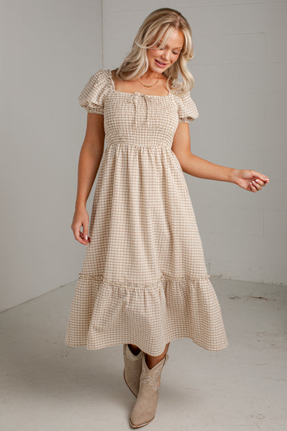 A woman modeling a beige and white gingham tiered midi dress with puff sleeves and a smocked bodice, paired with beige ankle boots, in a minimalist studio setting.