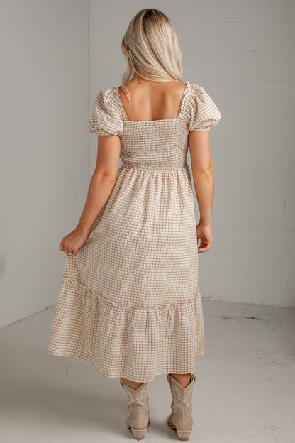 A woman modeling a beige and white gingham tiered midi dress with puff sleeves and a smocked bodice, paired with beige ankle boots, in a minimalist studio setting.