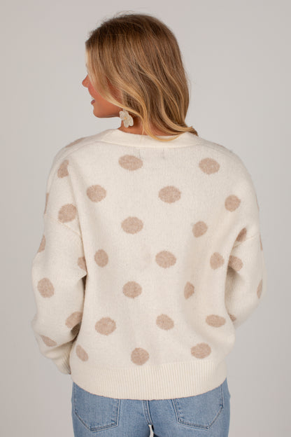 A woman viewed from the back wearing a cream-colored knit sweater with beige polka dots, paired with light blue jeans, against a solid light gray background.