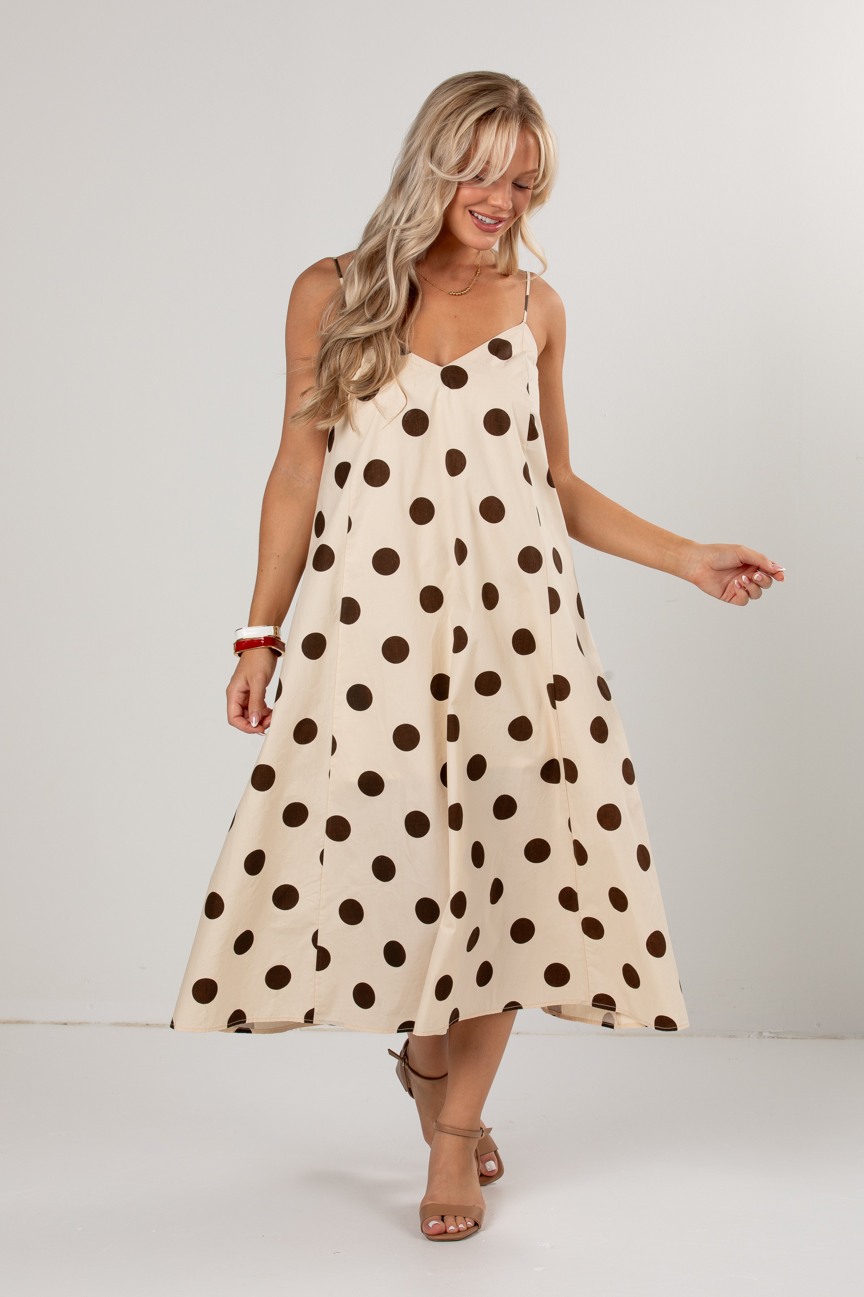 A woman modeling a sleeveless midi dress with a cream base and dark brown polka dots, set against a plain light gray studio background.