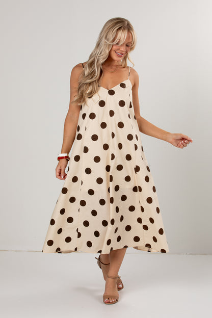 A woman modeling a sleeveless midi dress with a cream base and dark brown polka dots, set against a plain light gray studio background.