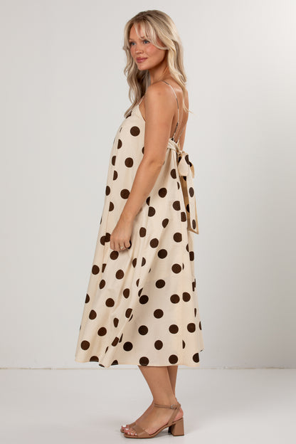 A woman modeling a sleeveless cream midi dress with large dark brown polka dots, shown from a side-back angle against a plain light gray studio background.