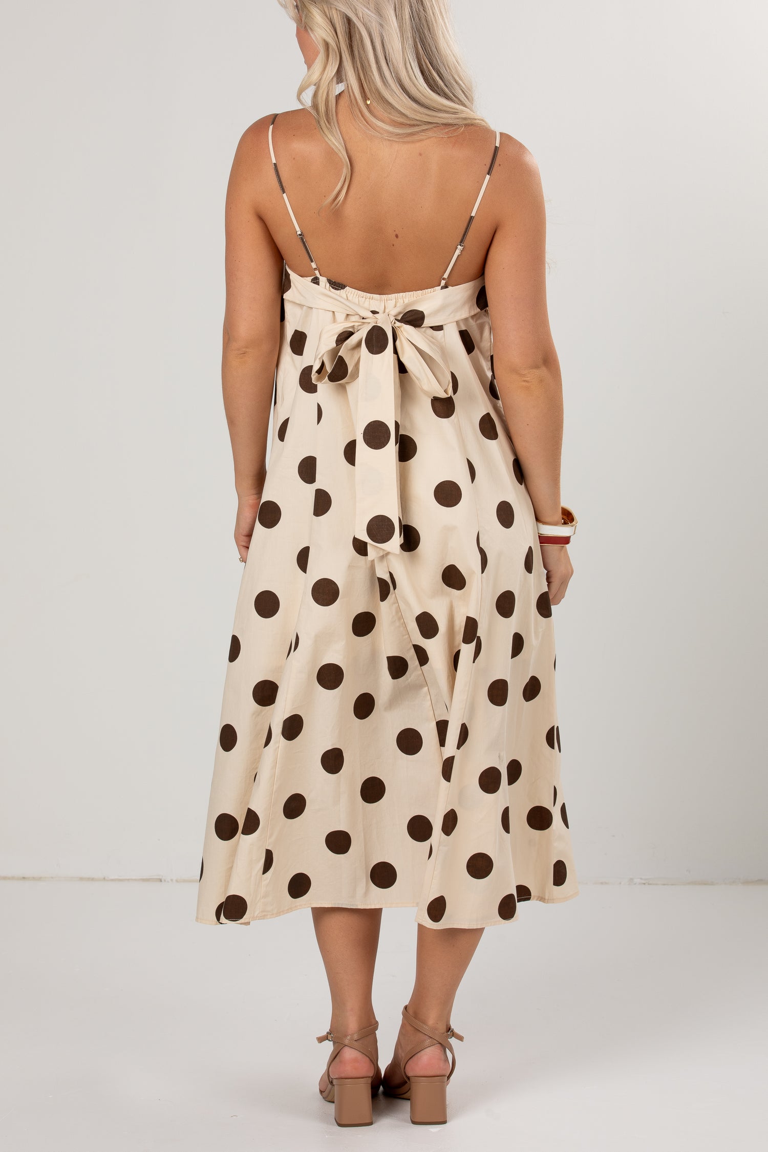 A woman viewed from the back wearing a cream midi dress with dark brown polka dots and thin shoulder straps, paired with brown heeled sandals against a plain light gray studio background.