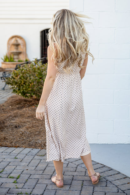 Woman in a polka dot dress walking on a paved path with a white wall and plant in the background.