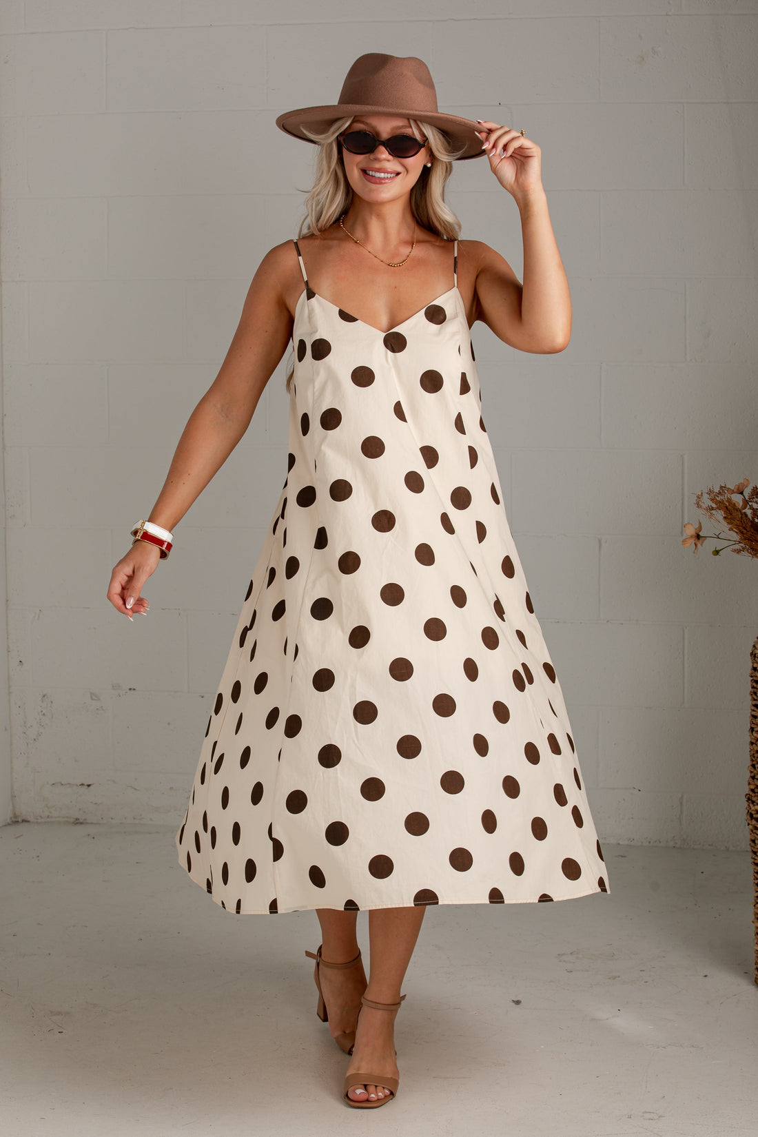 A woman models a cream-colored midi slip dress with brown polka dots, styled with a brown wide-brim hat, sunglasses, and tan heeled sandals in a bright studio setting.