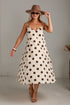 A woman models a cream-colored midi slip dress with brown polka dots, styled with a brown wide-brim hat, sunglasses, and tan heeled sandals in a bright studio setting.