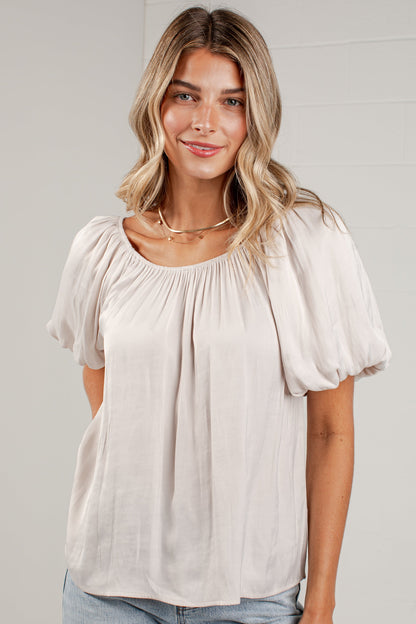 A woman wearing a light beige short-sleeve blouse with a gathered, balloon-style neckline and light-wash blue jeans, standing against a light gray paneled wall.