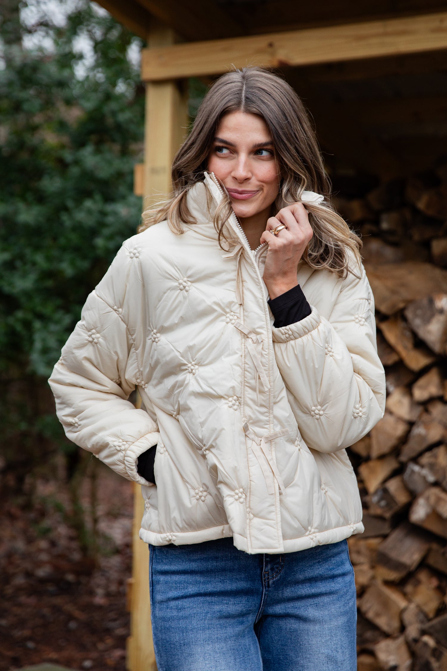 Woman wearing a cream quilted jacket standing in front of stacked firewood.
