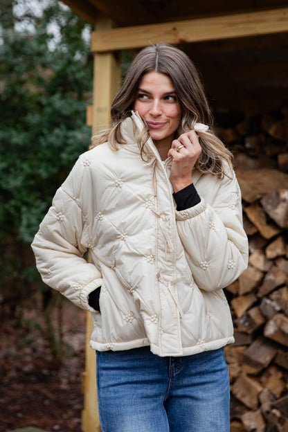 Woman wearing a cream quilted jacket standing in front of stacked firewood.