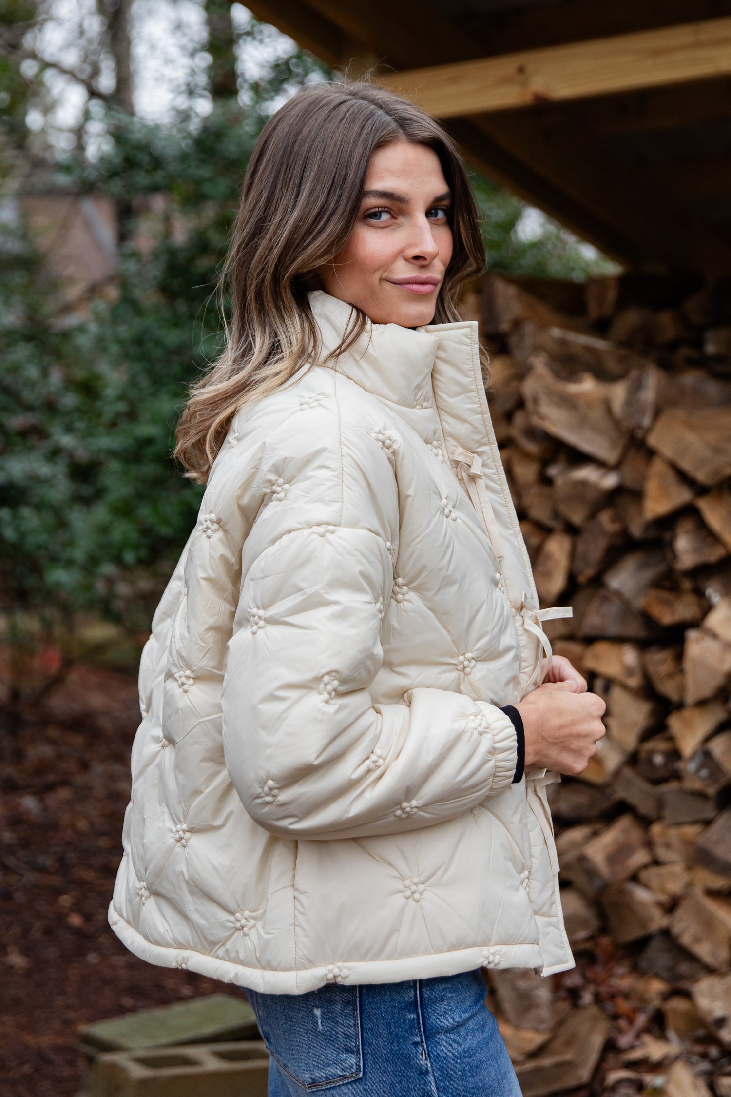 Woman wearing a white quilted jacket standing in front of stacked firewood.