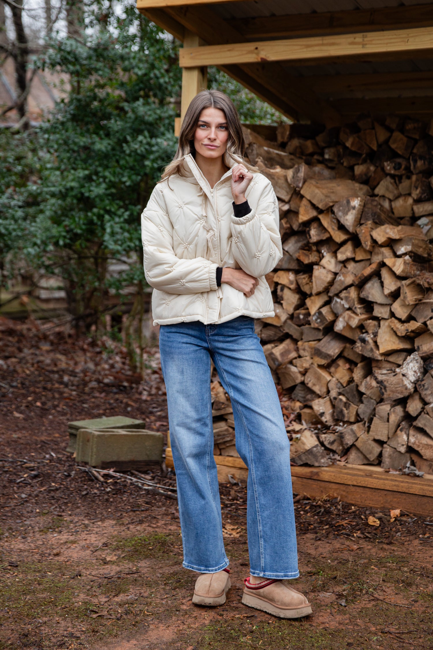 Person wearing a cream puffer jacket and blue jeans standing in front of stacked firewood.